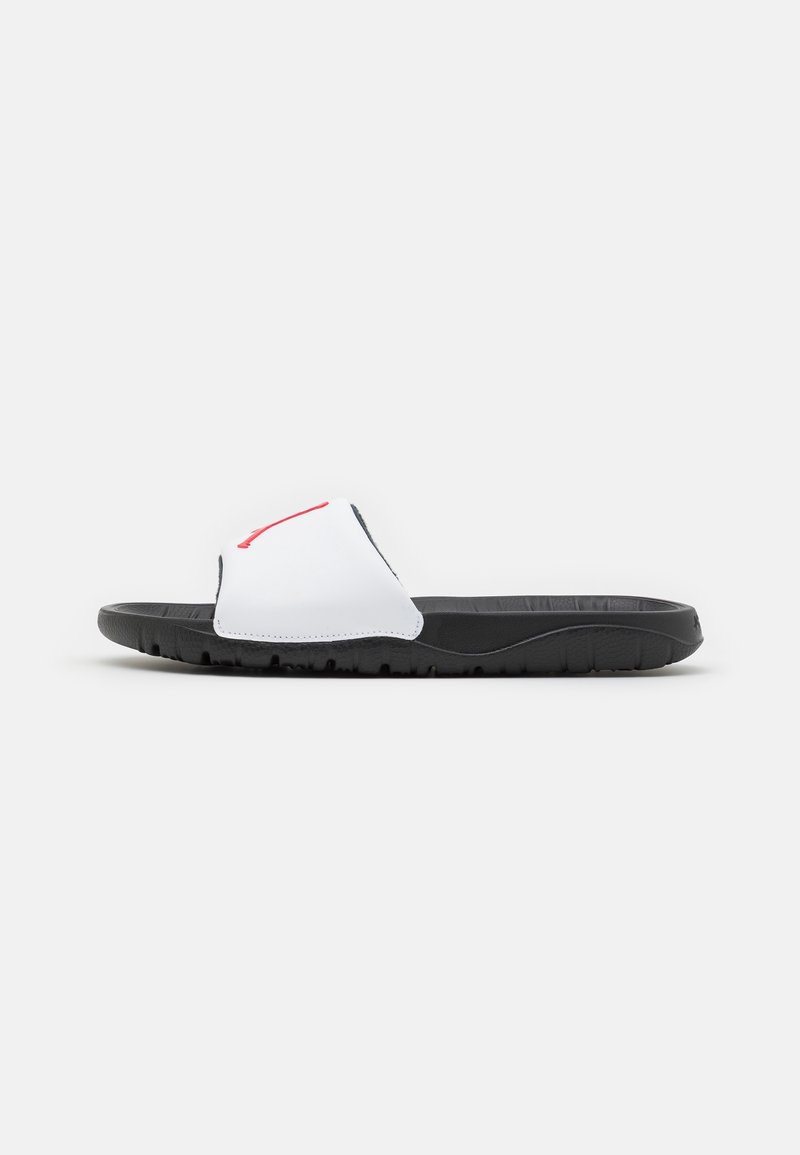 Sandale noire à semelle texturée et bride blanche avec un élément de design rouge sur un fond clair uni.