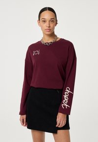 Burgunderfarbendes Langarmshirt mit weißem grafischem Text auf dem Ärmel, kombiniert mit einem schwarzen Cordrock. Hat einen runden Ausschnitt und eine lässige Passform.