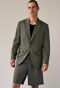 Olijfgroen tailleurjack met notches op de revers, knoopsluiting aan de voorkant en twee zijzakken, gecombineerd met een bijpassende knielange broek in een vergelijkbare tint.