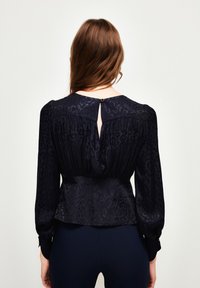 Marinebluse mit strukturiertem Muster, gerafftem Bund, langen Ärmeln und einem Schlüsselloch-Rückenverschluss, der mit einem goldfarbenen Knopf gesichert ist. Glatte Stoffqualität.