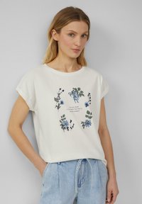 T-shirt en coton couleur crème avec des manches roulées, arborant des graphismes floraux en bleu et vert, et un texte motivant au centre.