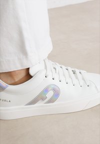 Sneaker bianco in pelle con logo iridescente, punta tonda e suola piatta. Presenta lacci bianchi e un design minimalista. Adatta per un abbigliamento informale.