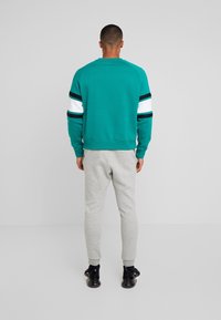 Turkos sweatshirt med svarta och vita ränder på ärmarna, tillsammans med gråa sweatpants och svarta sneakers; enkel bakgrund.