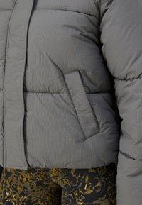 Šedá zateplená bunda s detaily bočních kapes, nošená přes legíny s abstraktním vzorem v žluté, černé a modré barvě.