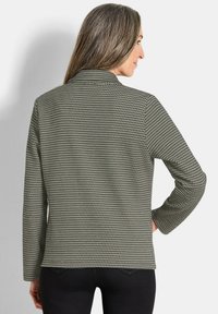 Houndstooth-gestreepte jas in olijfgroen en zwart, met lange mouwen, kraag en een zachte textuur. Ontworpen met een rechte snit en een iets getailleerde stijl.