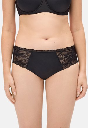 Mujer con sujetador negro y bragas negras a juego con paneles de encaje, de pie con las manos relajadas a los lados.