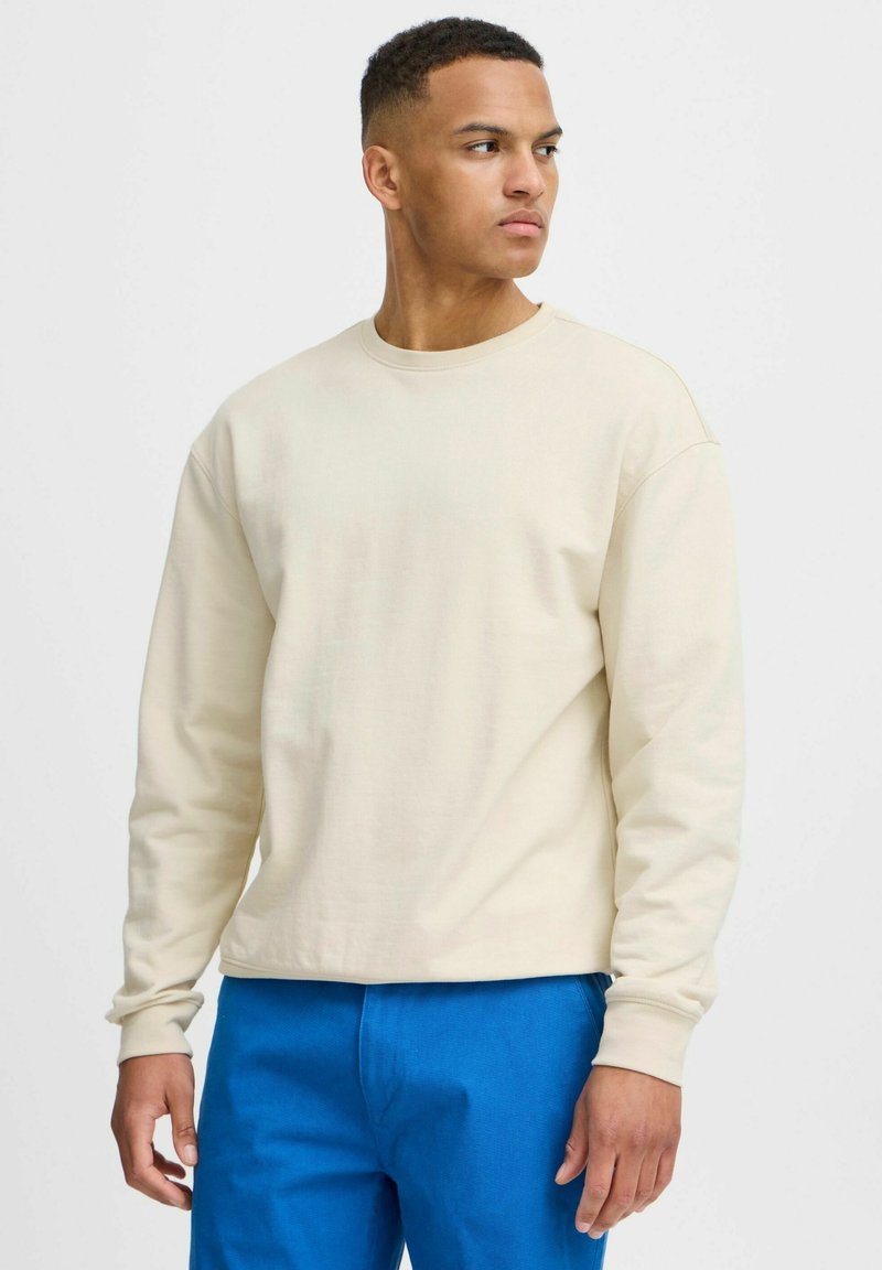 Sweatshirt en coton beige avec un col rond, coupe ample et manches longues, associé à un pantalon bleu vif. Texture simple et douce.
