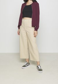 Burgundská bomber bunda na zip, černý crop top, béžové kalhoty s širokými nohavicemi, zelené a bílé tenisky s klasickým designem.