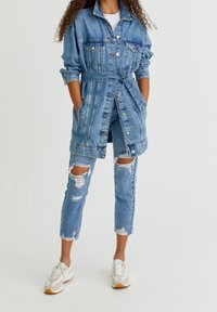 Veste en jean avec une taille ceinturée, deux poches poitrine, et un jean déchiré usé jusqu'aux genoux. Baskets blanches avec des accents gris et marron.