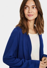 Blauwe gebreide cardigan met een open voorkant en lange mouwen, gedragen over een crèmekleurig top. Glad textuur en lichtgewicht ontwerp.