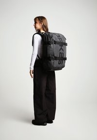 Mochila impermeable negra con un logo prominente, correas ajustables y acentos de hebilla. Diseño rectangular resistente con textura mate.