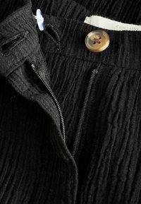 Material textil negru cu o textură, dotat cu fermoar, închidere cu nasture și detalii de cusătură. Prezintă un eticheta din fibră naturală vizibilă în partea de sus.
