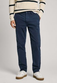 Man med marinblå chinos, krämfärgad stickad tröja med svarta ränder och vita sneakers med gummisulor, med händerna i fickorna, stående på grått golv.