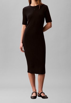 Femme portant une robe midi noire ajustée à manches courtes et des chaussures plates Mary Jane noires, debout devant un fond gris uni.