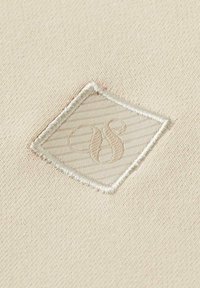 Tessuto beige con un patch quadrato cucito che presenta iniziali in rilievo. La superficie è liscia con sottili linee diagonali sul patch.