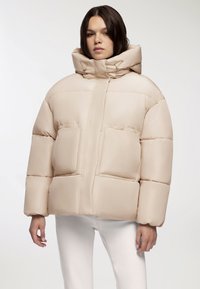 Beżowa kurtka puchowa z dużym kapturem, poziomym pikowaniem, dwoma przednimi kieszeniami i gładką, błyszczącą teksturą.