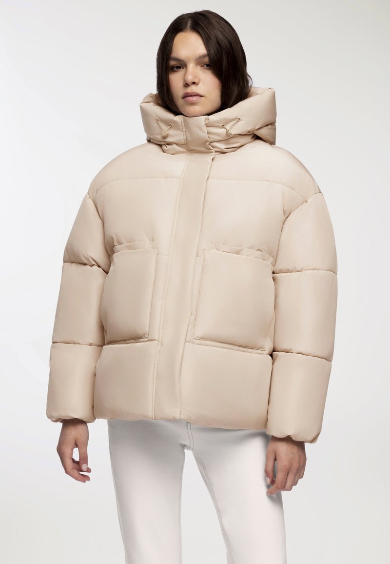 Beżowa kurtka puchowa z dużym kapturem, poziomym pikowaniem, dwoma przednimi kieszeniami i gładką, błyszczącą teksturą.