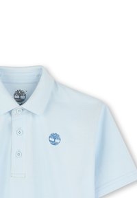 Polo shirt bleu clair en tissu texturé, avec un col, trois boutons et un logo d'arbre bleu sur le côté gauche de la poitrine.