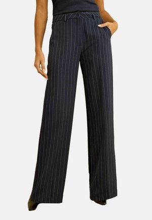 Femme portant un pantalon large noir à pinces et un haut noir, une main dans la poche, debout devant un fond uni.