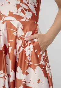 Satin-Kleid in Rostrot mit einem weißen Blumenmuster; es verfügt über eine seitliche Tasche und Falten für zusätzliches Volumen. Glatte Textur und figurnahes Design.