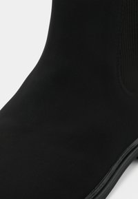 Gros plan sur une bottine noire en matériau lisse avec un panneau latéral élastique côtelé, mettant en valeur la tige de la chaussure et le bord de la semelle.