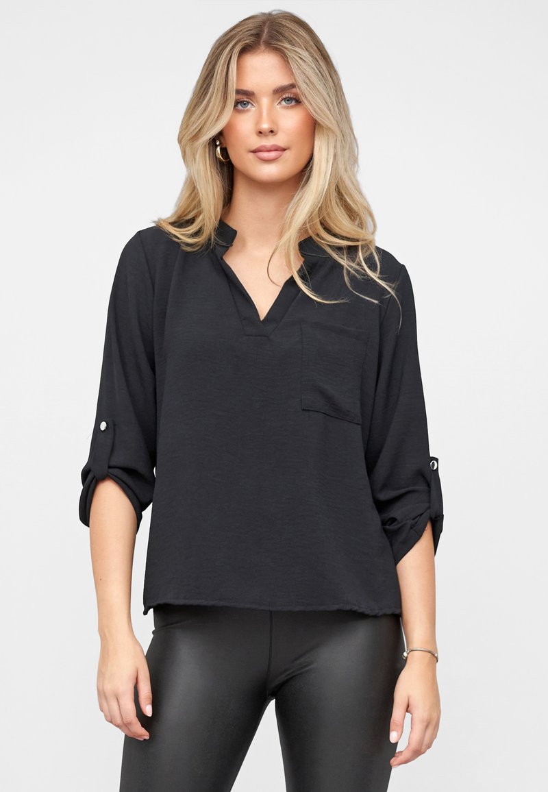 Schwarze Bluse mit V-Ausschnitt, langen Ärmeln, die bis zu den Ellbogen hochgekrempelt sind, strukturierter Stoff und Brusttasche. Kombiniert mit engen schwarzen Leggings.