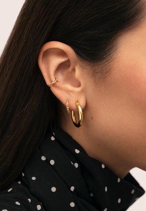 Gros plan sur l'oreille d'une femme avec trois boucles d'oreilles en or : un anneau lisse, un petit anneau orné de pierres précieuses, et une manchette d'oreille torsadée.