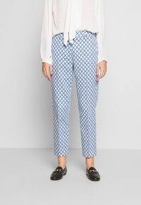 Pantalon bleu à motifs avec un design en losanges, fabriqué en tissu léger. Coupe droite, taille mi-haute, porté avec un chemisier blanc.