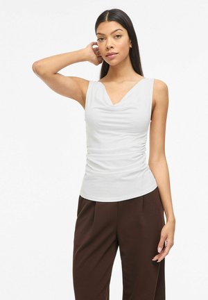 Femme aux longs cheveux noirs portant un haut blanc sans manches froncé et un pantalon large marron, debout avec une main sur la nuque.