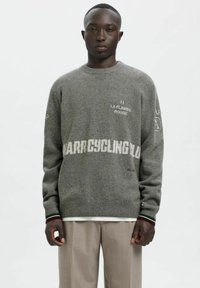 Pelēks adīts džemperis ar apaļu izgriezumu, kam ir balts un sarkans izšūts teksts. Ribotas aproces zaļā un vīnsarkanā krāsā.