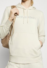 Sudadera con capucha de color crema con un bolsillo tipo canguro y un sutil logo de "Calvin Klein". Acompañada de una correa de mochila marrón con textura.