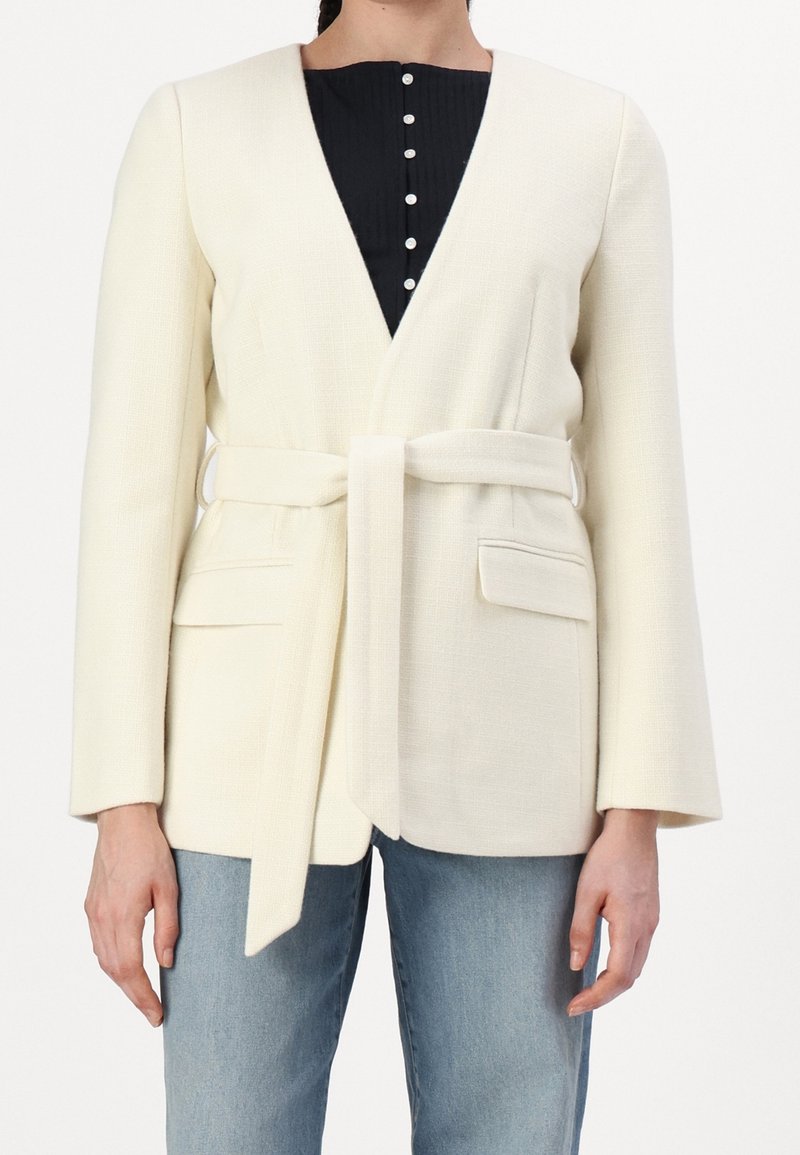Femme portant un blazer crème ceinturé sur un top noir boutonné et un jean bleu, avec des poches à rabat à l'avant du blazer.
