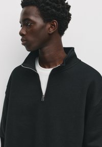 Črna zip-up jopica z visokim ovratnikom in rebrasto teksturo, nosena čez belo majico. Detajl zadrge je izpostavljen.