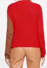Pull en tricot rouge à côtes avec des manches texturées marron. Il présente un col rond et une coupe décontractée. Porté avec un pantalon en cuir beige.