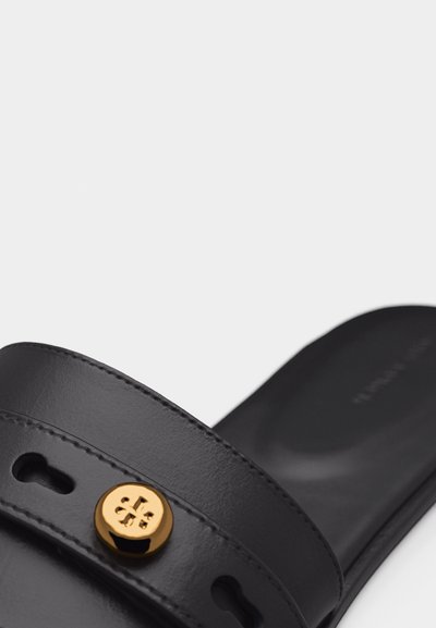 Μαύρο δερμάτινο σανδάλι με φαρδιά λωρίδα που διαθέτει χρυσό κυκλικό έμβλημα και οπές σε σχήμα κλειδαρότρυπας.