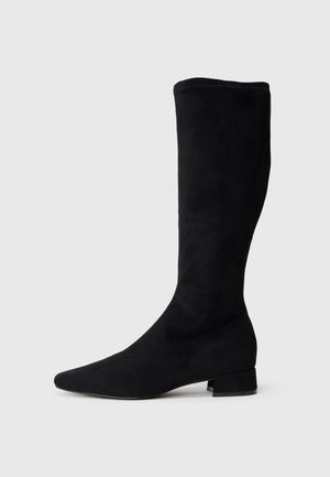 Bota negra hasta la rodilla hecha de ante, con punta afilada y pequeño tacón cuadrado, que presenta una textura suave y una silueta ajustada.