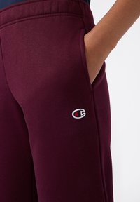 Ręka osoby w kieszeni bordowych dresów z logo Champion na udzie, na białym tle.