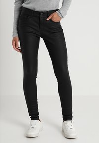 Černé skinny džíny s hladkým, elastickým materiálem, zapínáním na knoflíky a zip, a předními kapsami. V kombinaci s bílými teniskami a šedým topem.
