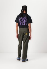 Schwarzes T-Shirt mit lila Grafikdesign, olivgrüne Hose, blaue Sneakers mit weißen Streifen und einem gelben Etikett auf der Gesäßtasche.