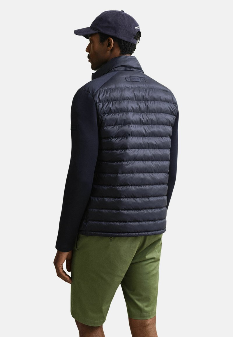 Mann trägt eine marineblaue gesteppte Jacke, eine marineblaue Kappe und grüne Shorts, gezeigt von hinten vor einem einfarbigen hellen Hintergrund.