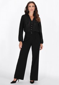 Musta pitkähihaisen v-aukkoinen pusero, jossa on napit edessä, yhdistetty korkeavyötäröisiin mustiin leveälahkeisiin housuihin. Sileä kangas ja räätälöity istuvuus.