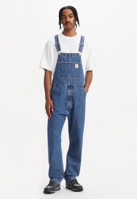 Salopette en denim bleu moyen avec bretelles réglables, poche frontale, coupe décontractée et ourlets retroussés, associée à un t-shirt blanc et des chaussures noires.