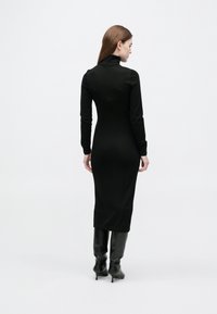 Γυναίκα με μακριά καστανά μαλλιά που φοράει μαύρο φόρεμα με μακρύ γιακά και μαύρες ψηλοτάκουνες μπότες μέχρι το γόνατο, στέκεται σε λευκό φόντο.