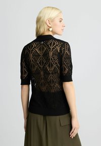 Maglione corto a maniche corte di colore nero, con un motivo in pizzo geometrico sulla schiena e una parte anteriore liscia. Presenta bordi a coste e una consistenza leggera.