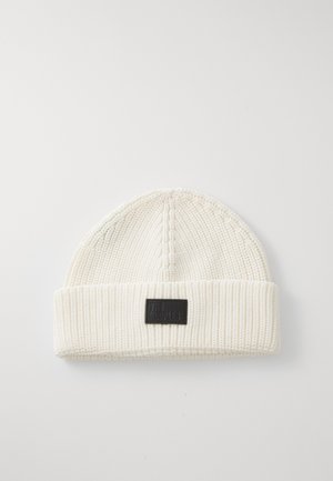 Cream-colored knitted beanie with folded brim and a black rectangular logo patch centered on the front.