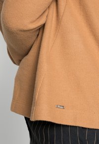 Beżowy sweter z dzianiny o żeberkowej teksturze. Posiada szczegóły w postaci bocznych rozcięć oraz małą metalową metkę z logo na dolnym brzegu. Zestawiony z spodniami w prążki.