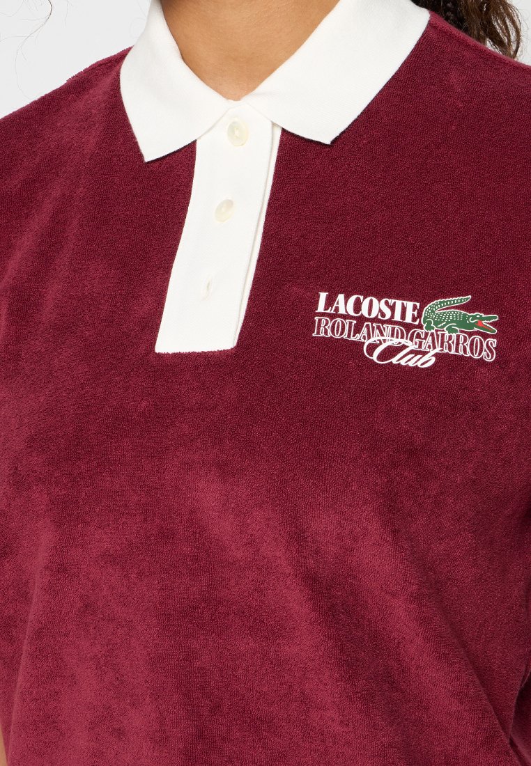 Polo en coton bordeaux avec un col blanc. Présente le logo brodé "LACOSTE ROLAND GARROS Club" et un tissu texturé. Patte à trois boutons.