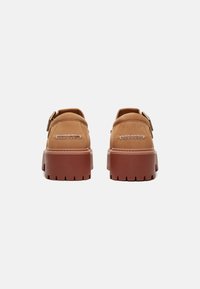 Sapatos de couro castanho com uma sola de borracha chunky, apresentando costuras e um detalhe de fivela lateral. Textura suave, forma arredondada e design robusto.