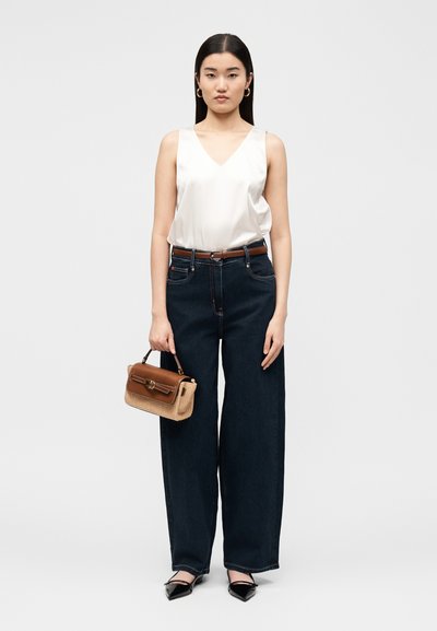 Γυναίκα που στέκεται φορώντας λευκό αμάνικο τοπ, σκούρα φαρδιά τζιν, μαύρα μυτερά παπούτσια, χρυσούς κρίκους για σκουλαρίκια και κρατώντας καφέ τσάντα.