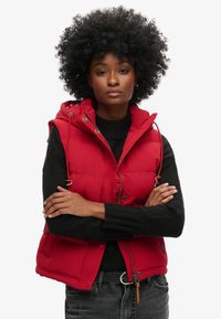 Chaleco puffer rojo con capucha, cremalleras y cierres a presión, y bolsillos laterales. Lleva puesto un suéter de punto negro y lo ha combinado con jeans oscuros.